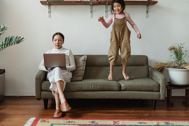 mom typing on laptop on couch with daughter jumping beside her